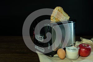 Black and silver electric micro oven with toast, fresh eggs, red apple and glass of milk on brown wooden and black background,