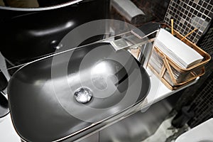 A black sink with a silver faucet next to an oval mirror and a shelf with hand paper towels. Close-up of an elegant