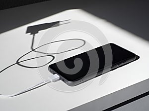 A black smartphone with a charging wire on a white table with shadows