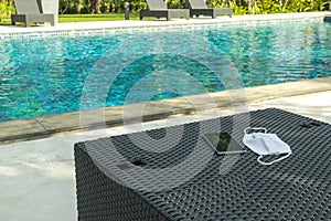 Black smartphone with white facial mask on weave chair nearly swimming pool.