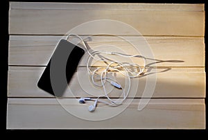 Black smartphone with white headphone is lying on wooden background. Modern technology connection. Mobile phone and earphone