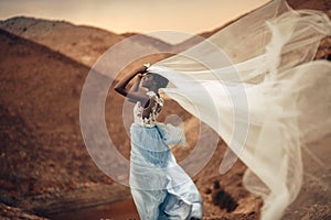 Black bride stands and holds waving bridal veil in her hands on background of beautiful landscape.