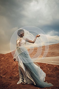 Black bride in waving long wedding dress and bridal veil stands on background of beautiful landscape.