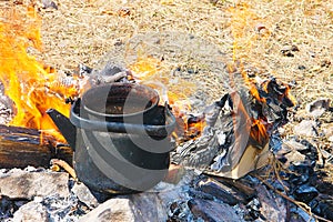 A black smoked teapot stands on fire surrounded by yellow flame tongues against the background of dry grass - tourist inventory