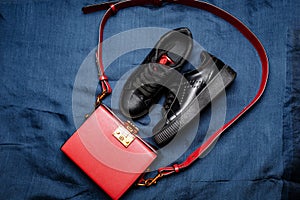 Black sneakers with red tongues and a red bag with a golden lock on a blue woven background