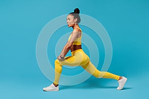 black sportswoman in yellow fitness outfit lunging on blue backdrop