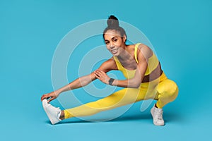 Black sportswoman in yellow fitwear stretching legs against blue background