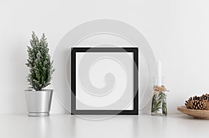Black square frame mockup with a cypress tree, candles and pine cones on a white table. Christmas decoration