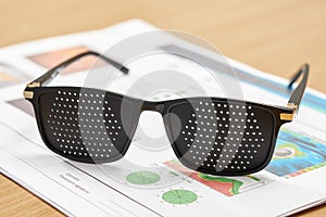 Black stenopeic glasses for eye muscle training placed on a table with blurred ophthalmic test results in a clinic setting.