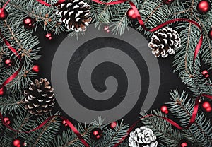 Black stone background with a frame of fir branches decorated with balls and red ribbon. Pine cones. Top view