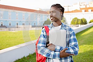 Black student guy hugs laptop standing with backpack near university