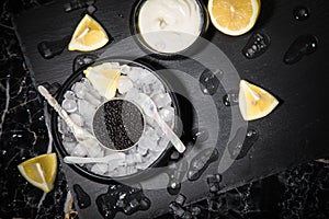 Black Sturgeon caviar on ice with pearl spoon, sour cream, lemon on slate plate and dark marble background