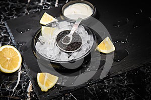 Black Sturgeon caviar on ice with pearl spoon, sour cream, lemon on slate plate and dark marble background