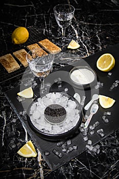 Black Sturgeon caviar on ice with pearl spoon, sour cream, toast, lemon and vodka in lead glass on slate plate and dark marble