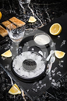 Black Sturgeon caviar on ice with pearl spoon, sour cream, toast, lemon and vodka in lead glass on slate plate and dark marble