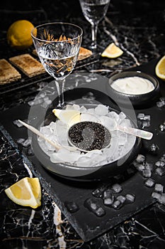 Black Sturgeon caviar on ice with pearl spoon, sour cream, toast, lemon and vodka in lead glass on slate plate and dark marble