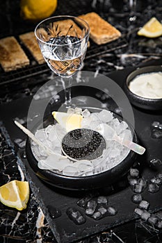 Black Sturgeon caviar on ice with pearl spoon, sour cream, toast, lemon and vodka in lead glass on slate plate and dark marble