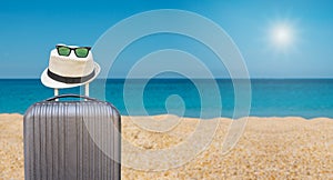 Black suitcase with hipster hat and sunglasses on tropical sea and sandy beach a in background. Summer holiday traveling design