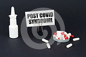 On a black surface are pills, a nasal spray and a paper sign with the inscription - Post covid syndrome