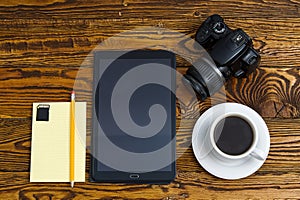 Black tablet computer on a old wooden table with a mug of coffee, camera and notebook with pencil