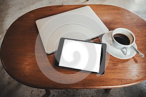 A black tablet pc with blank desktop white screen with laptop and coffee cup on wooden table