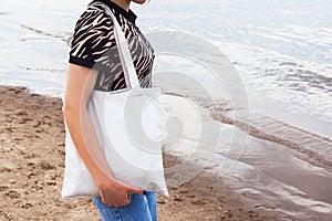 black tanned girl holding tote eco bag close up canvas fabric for mockup. young woman stands against of sand coast