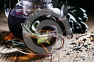 Black tea with fresh sage, glass cup and teapot, dark wood background, selective background
