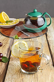 black tea in a glass cup with mint cherries and lemon on a wooden table and a teapot and lemon in a plate and mint