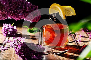 Black tea with lemon in a transparent glass cup, flying citrus slices and lilac branches and flowers