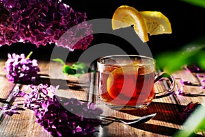 Black tea with lemon in a transparent glass cup, flying citrus slices and lilac branches and flowers