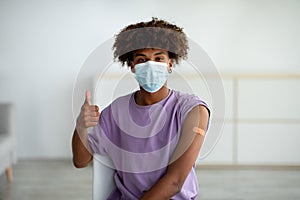 Black teen guy in face mask wearing plaster bandage on his arm after covid vaccine injection, showing thumb up at home