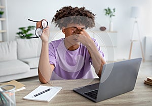 Black teen guy rubbing tired eyes, sitting at desk with laptop computer, exhausted from studying online