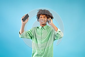 Black teen guy with smartphone and wireless headphones, listening to music, smiling to camera over blue background