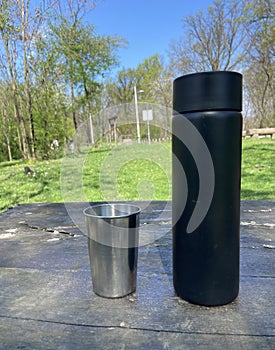 Black Thermos Bottle And Silver Metal Cup On Wooden Table In The Park