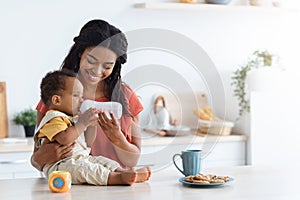 Black Toddler Boy Drinking Water From Bottle With Smiling Mom In Kitchen