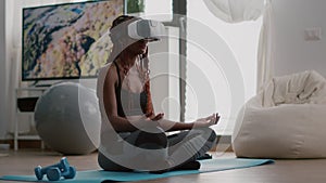 Black trainer woman wearing virtual reality headset while sitting on yoga map