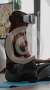 Black trainer woman wearing virtual reality headset while sitting on yoga map