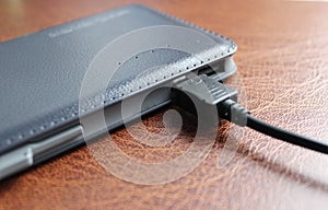 Black USB cable is connected to a discharged power bank with leather upholstery closeup angle view