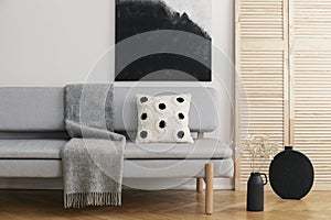 Black vase with cotton flower on the floor of bright trendy living room with grey sofa with pillow and warm blanket, real photo