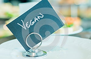 Black vegan sign with handwriting on a clip with blurred food in the background