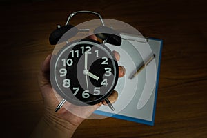 Black vintage alarm clock in hand with clipboard and blank white paper and pen, copy space for add your text, work on time or over