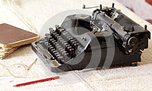 Black vintage metal type writer next to a pile of old note books, pair of eyeglasses and a pencil on a linen tablecloth