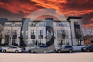 A black and white apartment building with trees in front and parked cars along the street with powerful red clouds at sunset