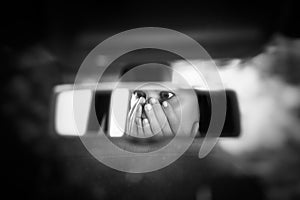 Black and white art picture of young woman with frightened eyes covering her face with hands in the car rear view mirror.