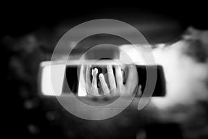 Black and white art picture of young woman with frightened eyes covering her face with hands in the car rear view mirror.