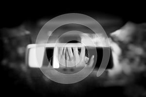 Black and white art picture of young woman with frightened eyes covering her face with hands in the car rear view mirror