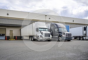 Black and white big rigs semi trucks with semi trailers loading cargo at warehouse dock with gates for each