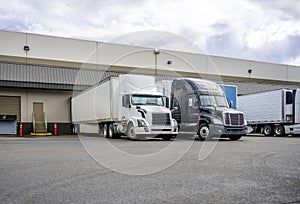 Black and white big rigs semi trucks with semi trailers loading cargo at warehouse dock with gates for each photo