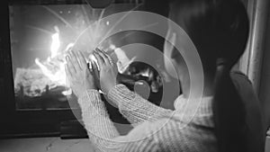 Black and white closeup image of woman sitting by the fireplace and warming her hands