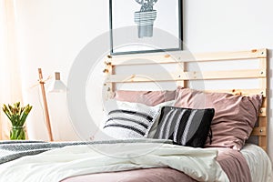 Black and white cushions placed on double bed with wooden bedhead in bright room interior with poster, fresh flowers and lamp